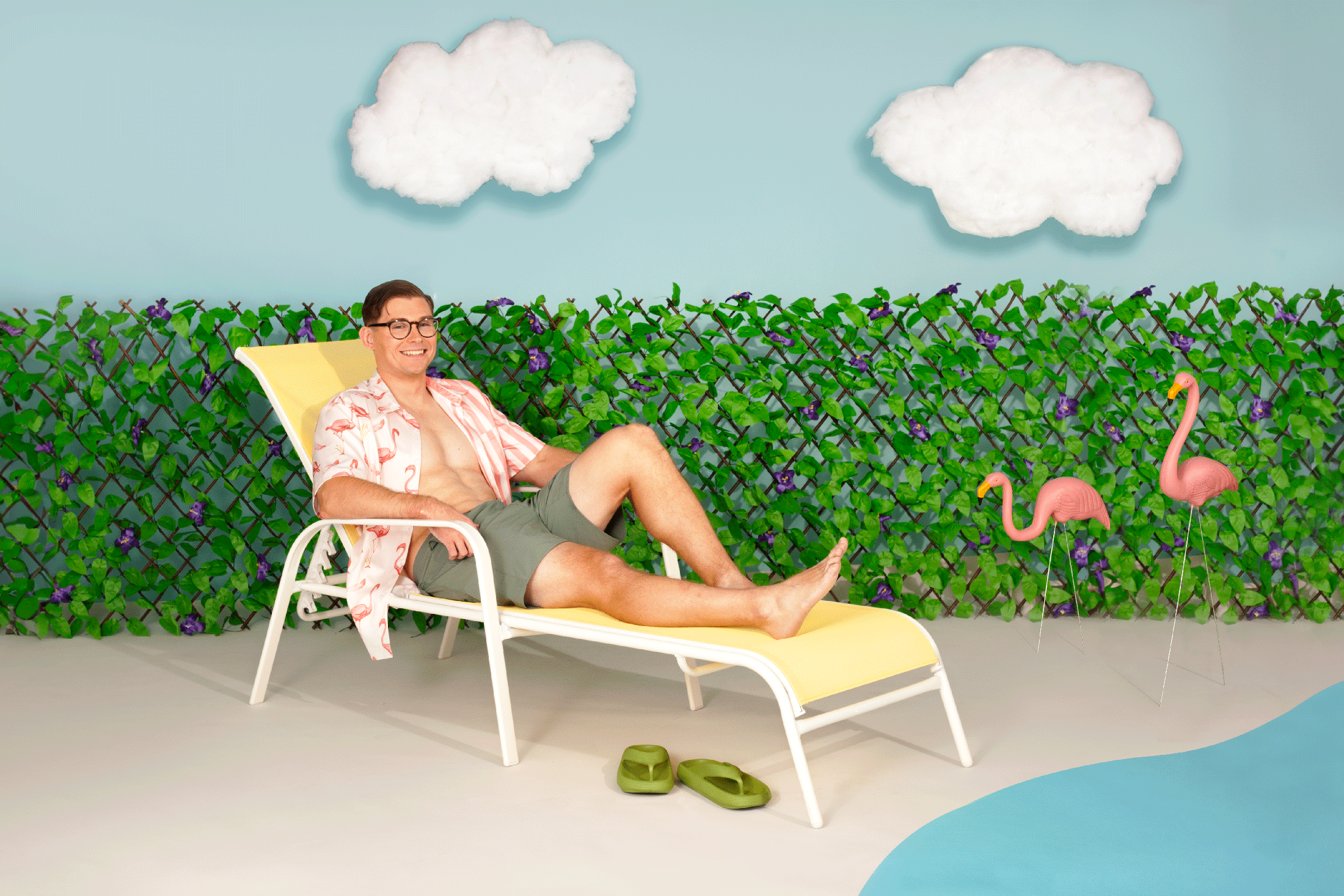 A man reclining on a lounge chair, smiling and relaxed in a staged summery setting with faux grass, a hibiscus-patterned shirt, and flamingo ornaments.