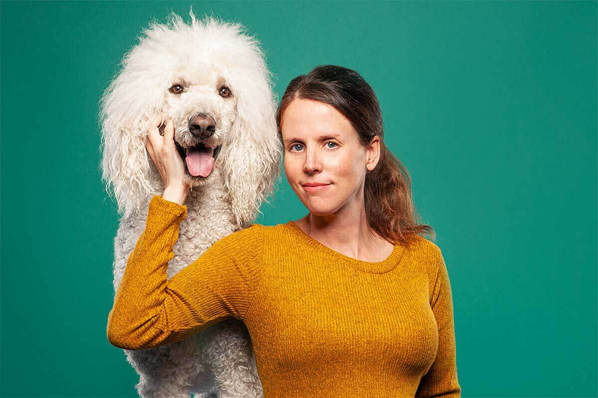 A woman in a mustard sweater smiling beside her fluffy white poodle against a teal background, showcasing a happy pet-owner bond in a studio portrait.