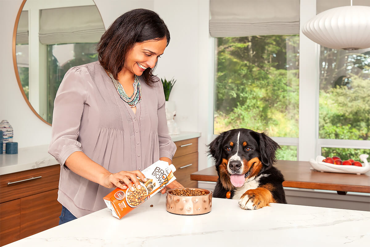 Smiling woman in a kitchen, pouring dog food topper into a bowl for an eager Bernese Mountain Dog.